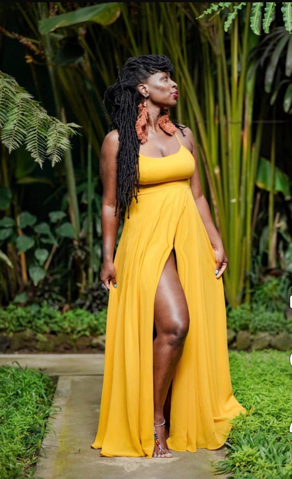 Black woman in a flowing yellow dress standing in lush tropical greenery, basking in natural sunlight